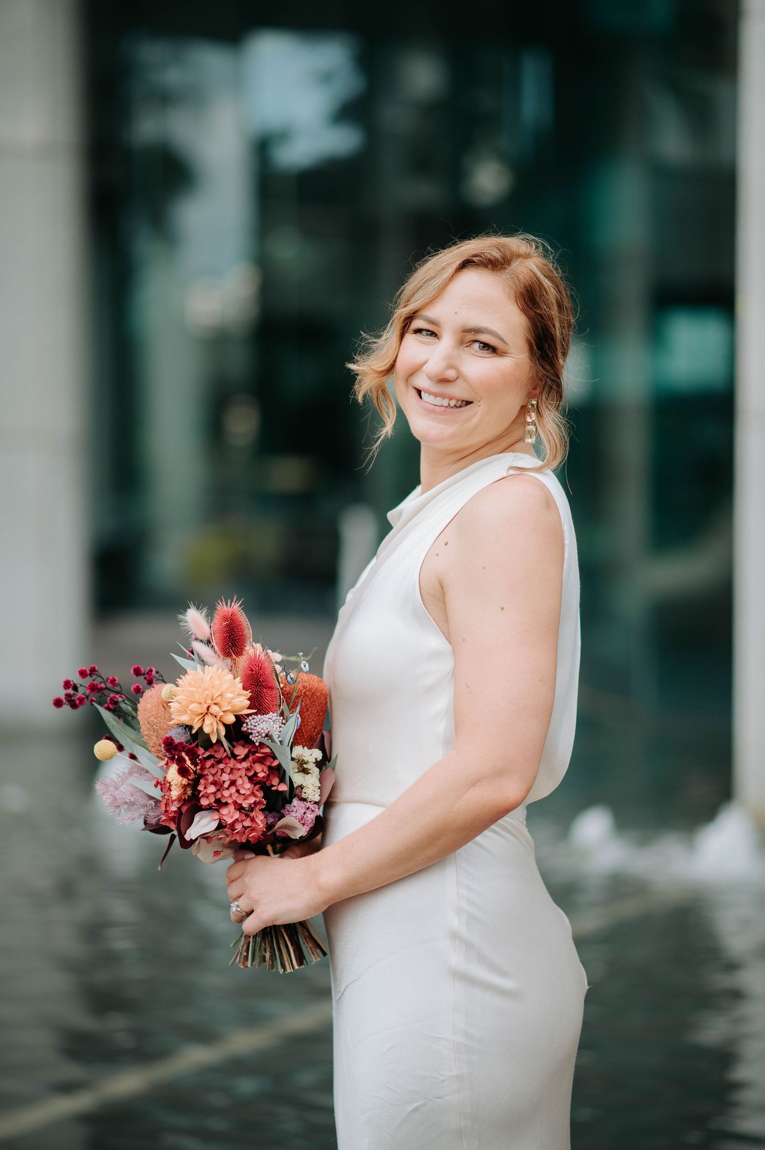 A bride in a sleek white dress holds a colorful bouquet while smiling at the camera.