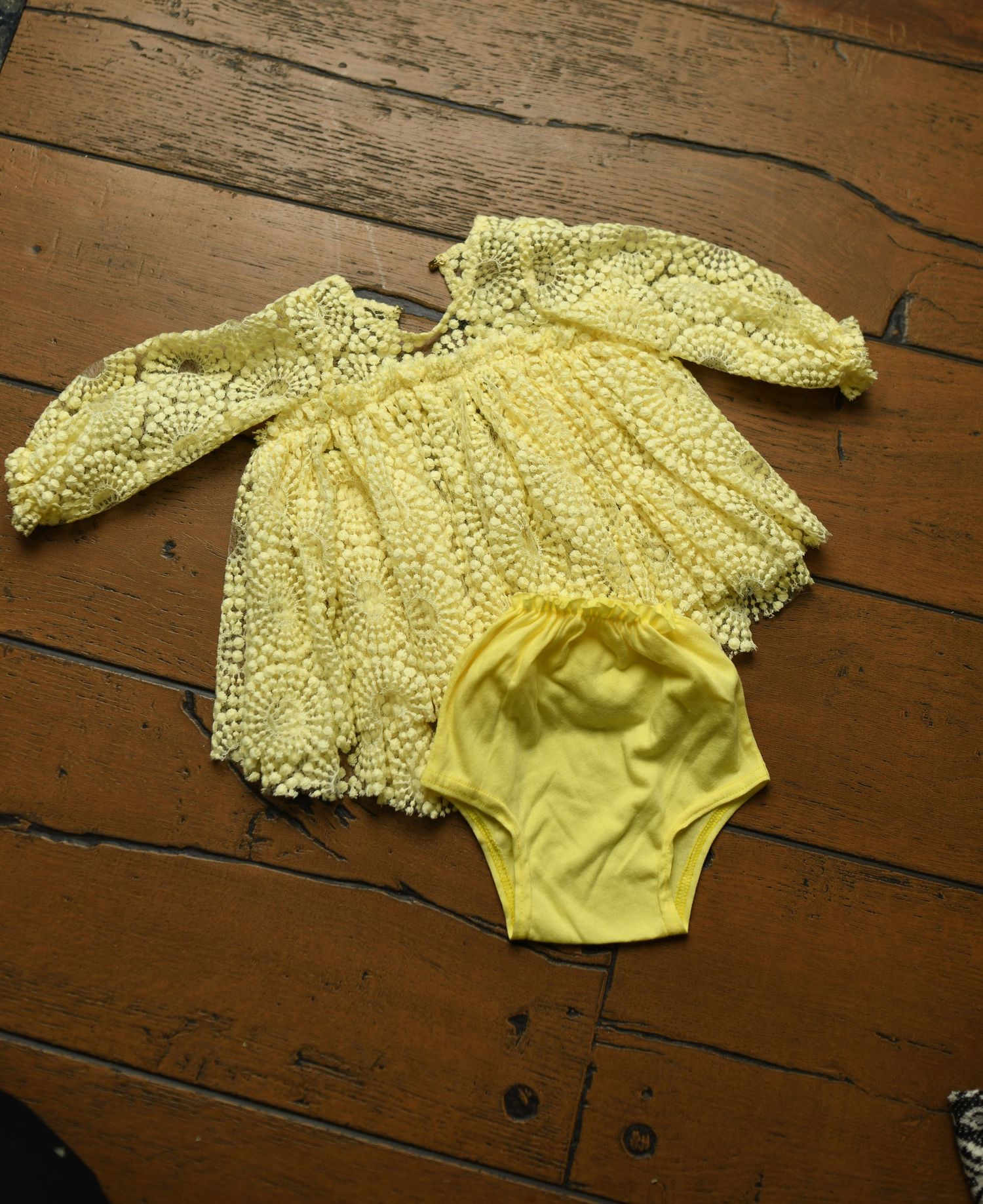 Yellow lace trimmed baby romper displayed on vintage wooden floorboards.