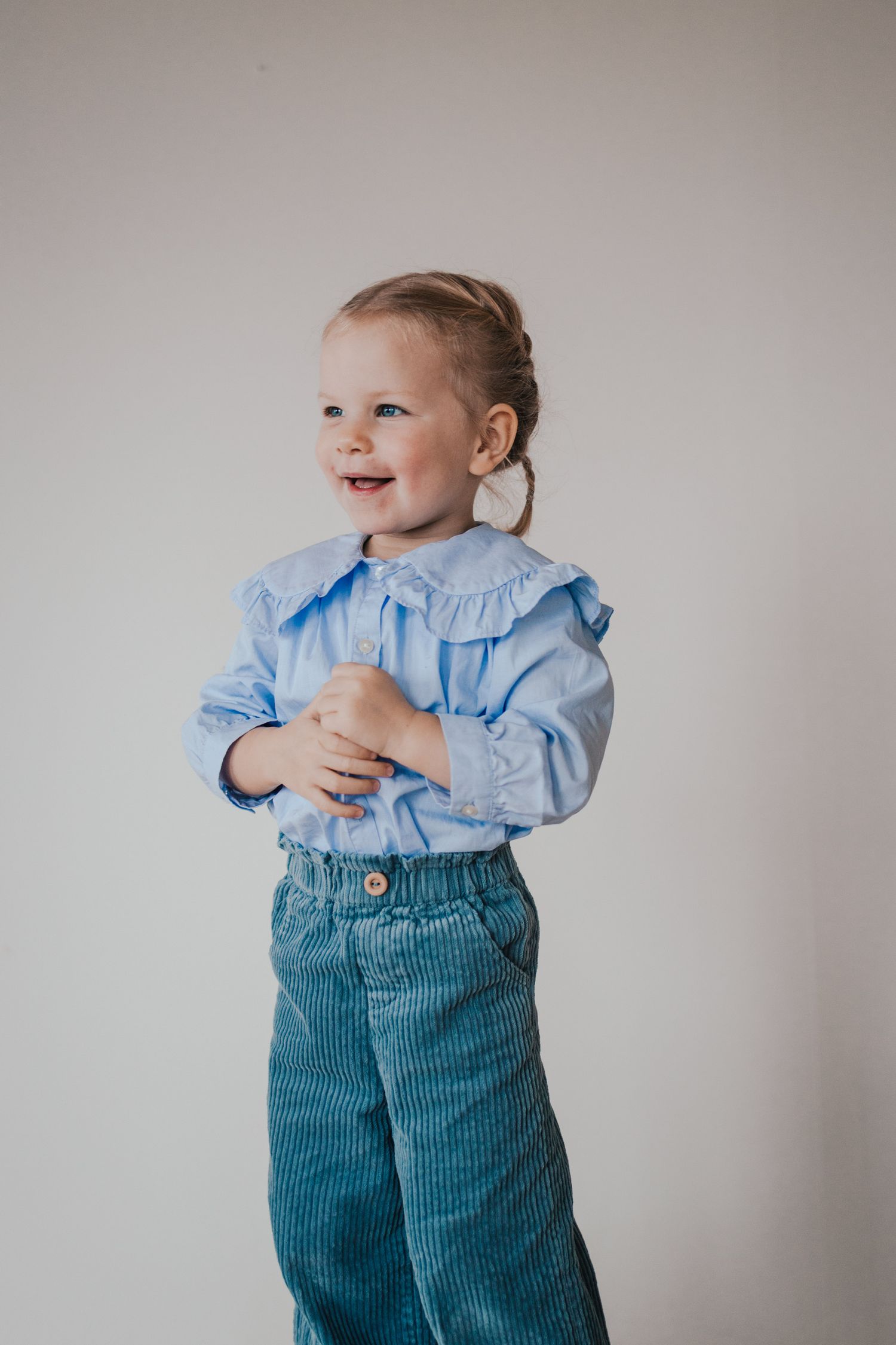 Small child wearing blue ruffled shirt and corduroy pants against neutral background.