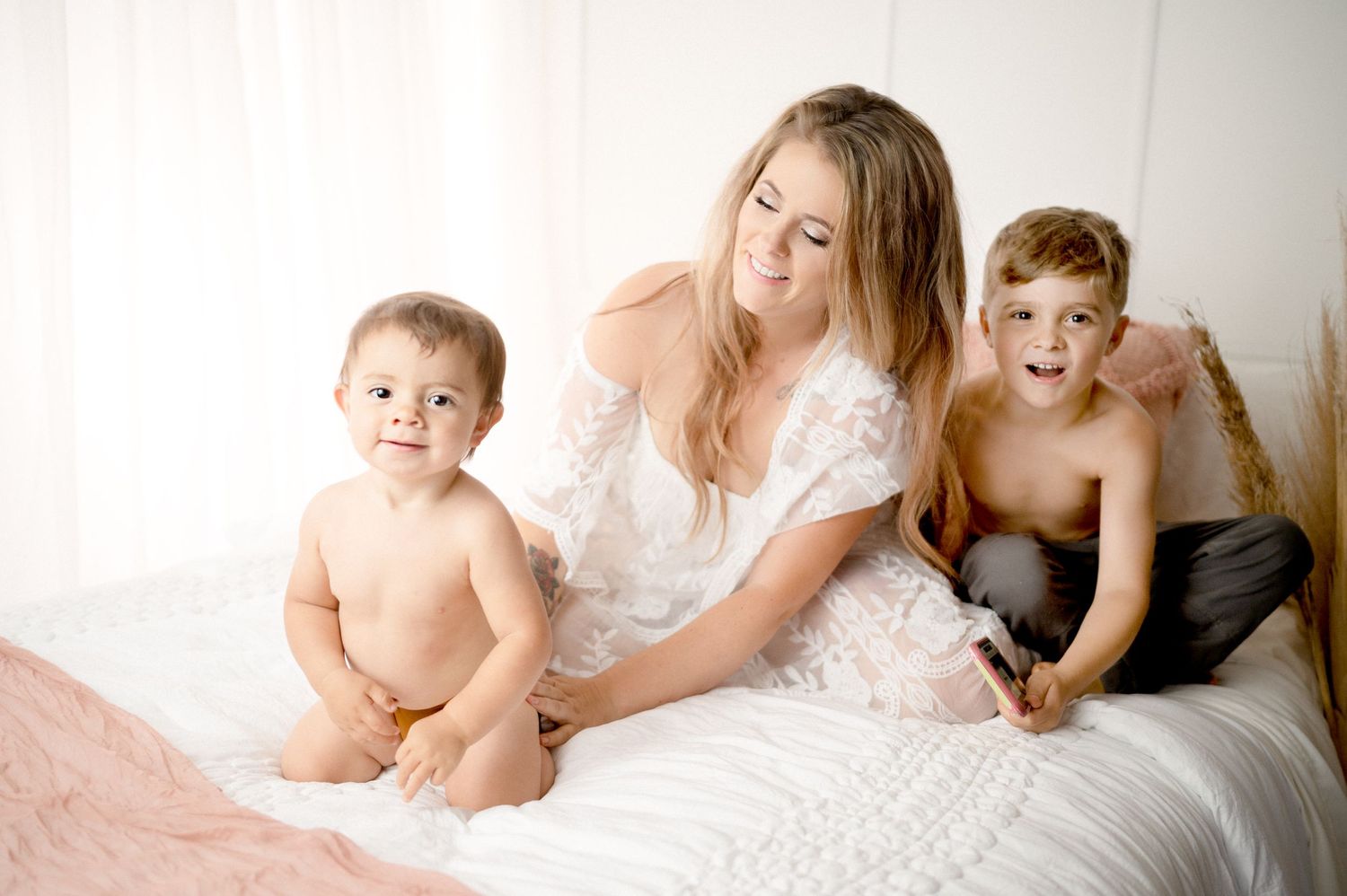 Mother in white dress plays with two young children on cozy bed.