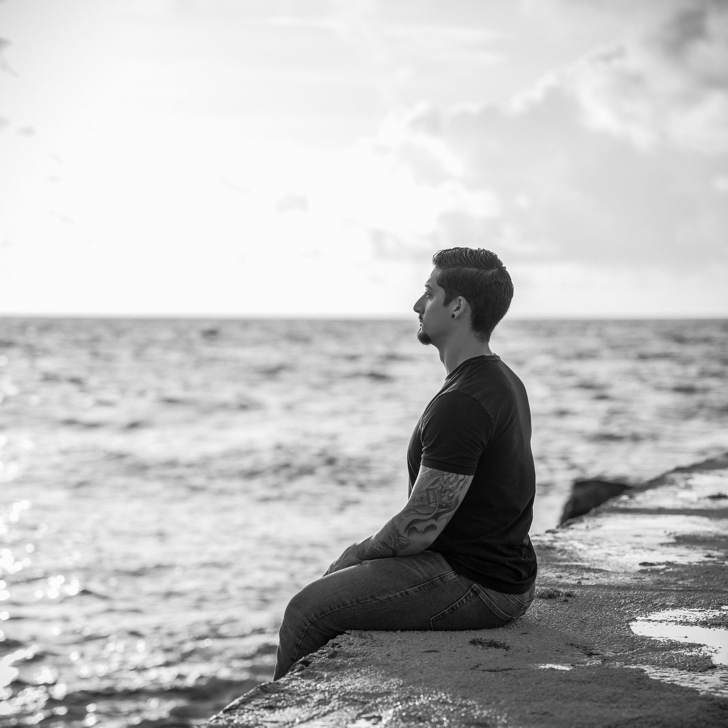 Person sitting alone on rocky shore looking out at ocean horizon in black and white.