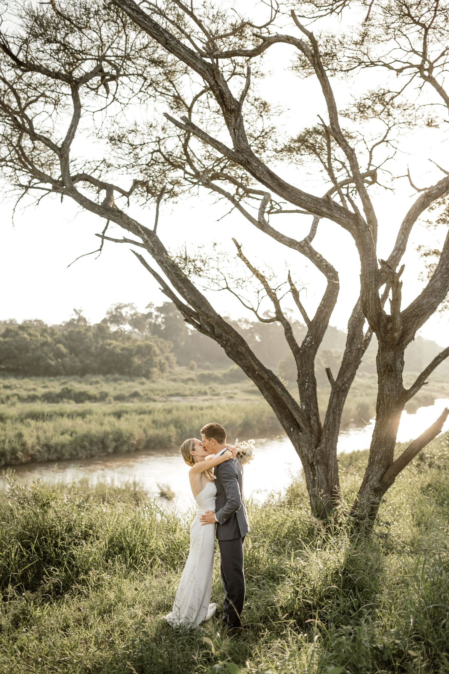 Bride and groom sharing a romantic kiss amidst tall grass, with a serene river and a lush tree in the background.