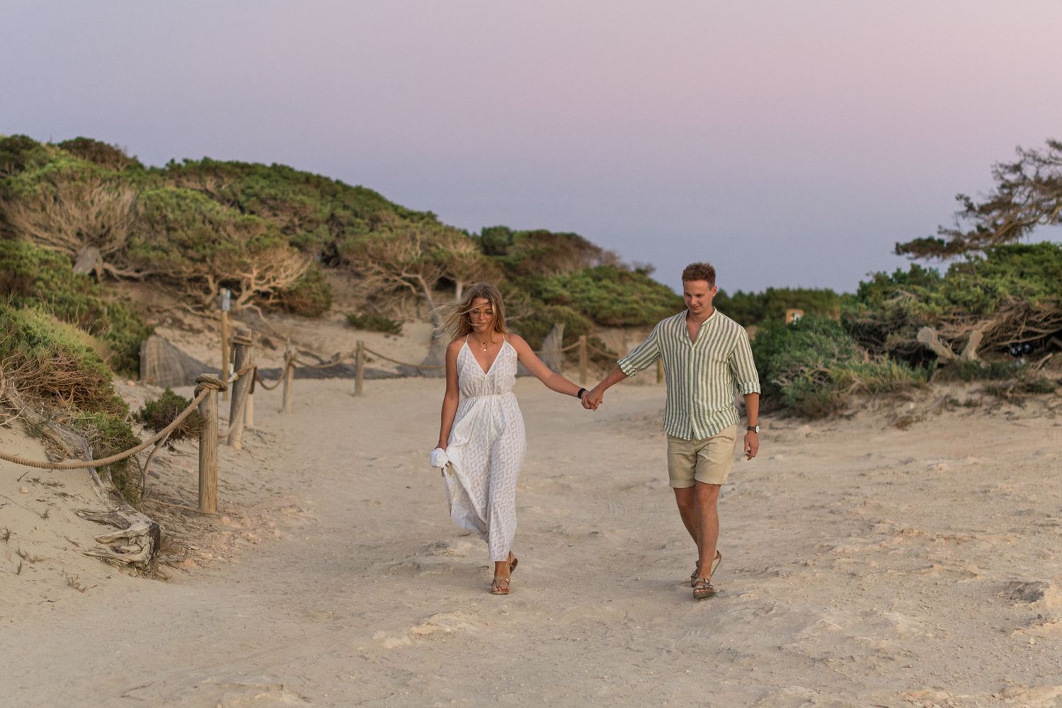 A couple hold hands and walk amongst the Ibiza sand dunes at sunset