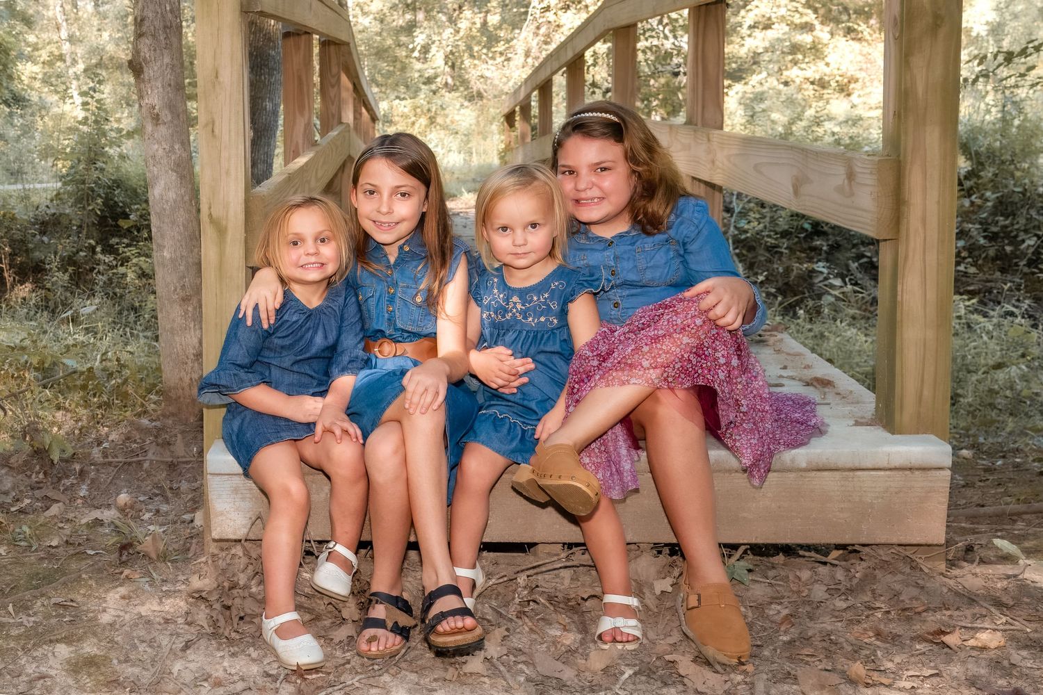 Four children in coordinated blue outfits sit together on a wooden bench outdoors.