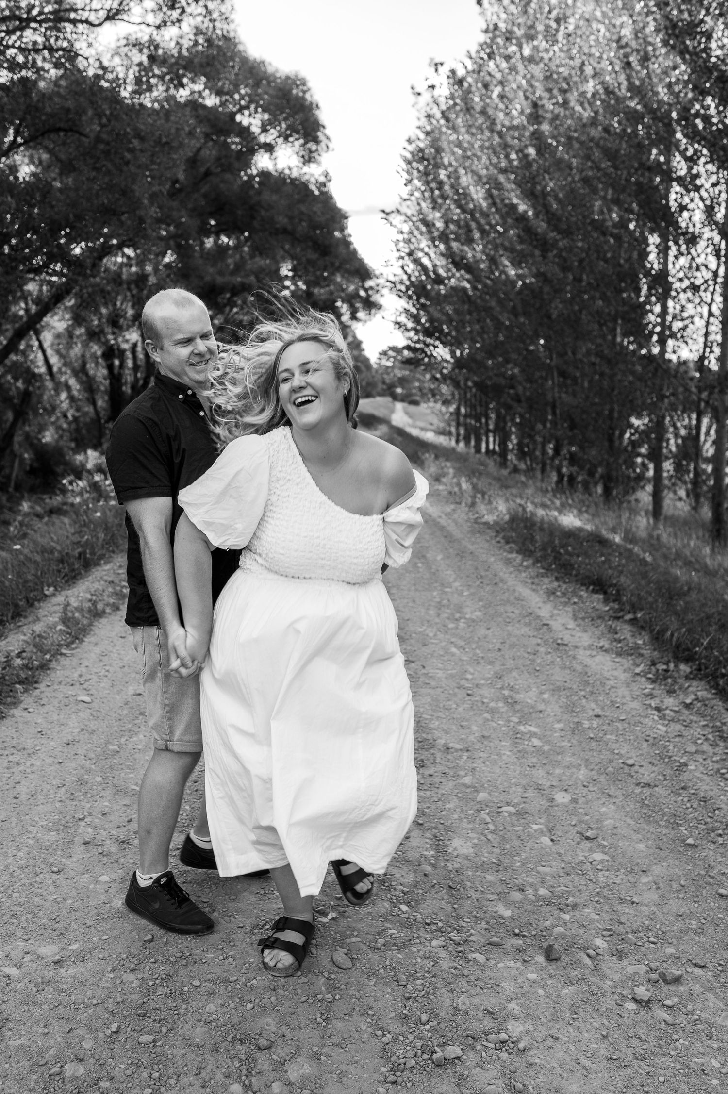 Couple laughing together on a tree-lined path in black and white photography.