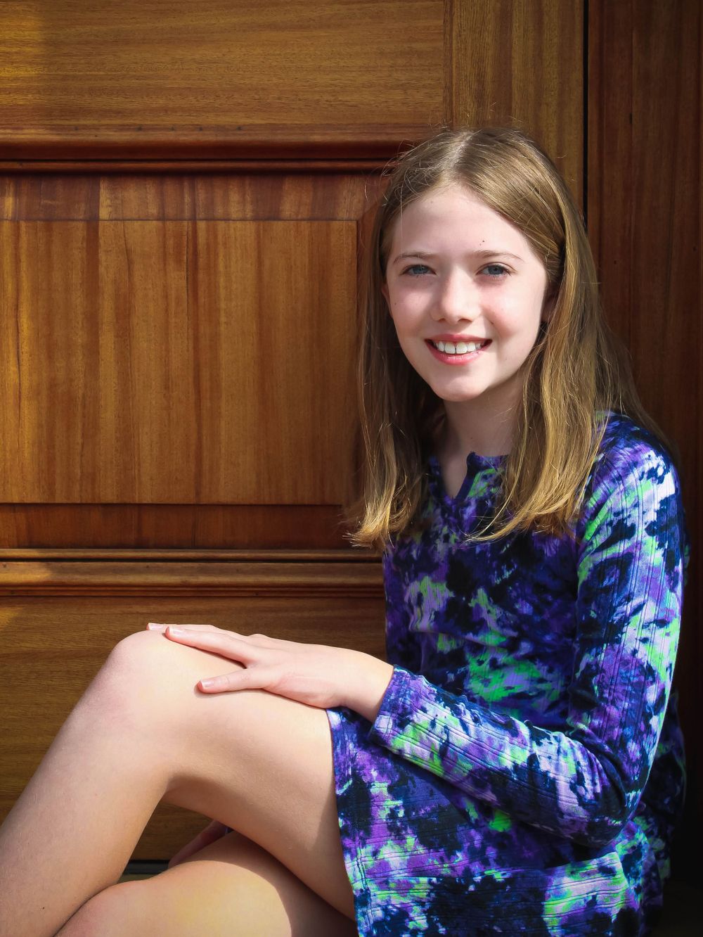 Someone seated against wood paneling wearing a blue and purple tie-dye dress.