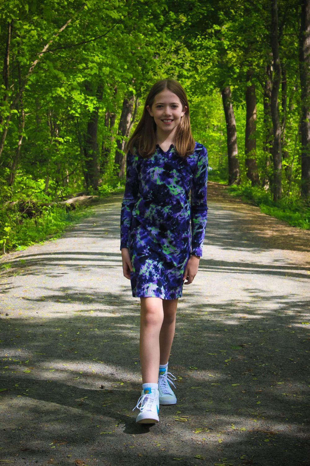 Someone walking on a tree-lined path wearing a blue tie-dye dress and sneakers.