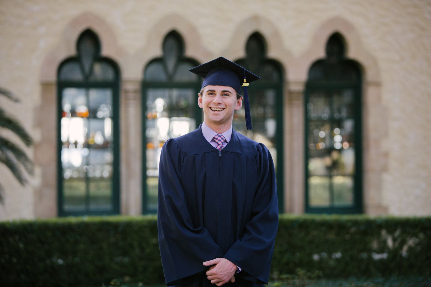 Graduate in cap and gown smiling in front of gothic style windows.