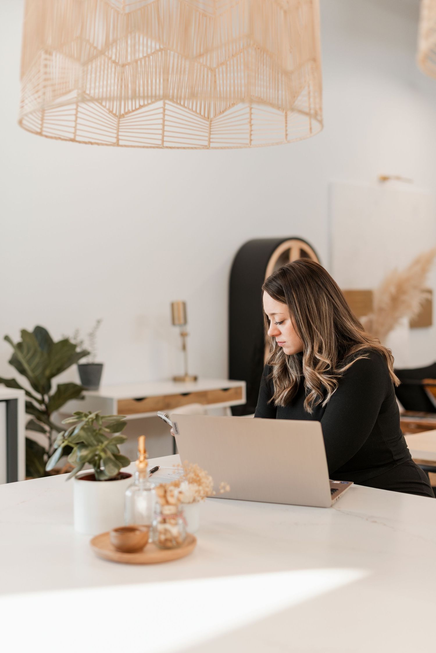 Woman working at minimalist white desk with laptop in sunlit modern office space.