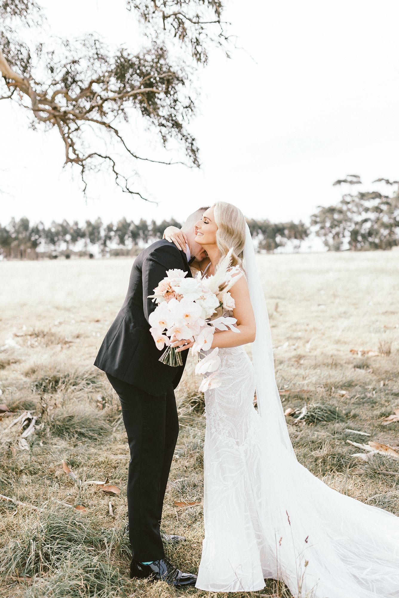 A romantic wedding embrace with blush pink bouquet under a gum tree branch.