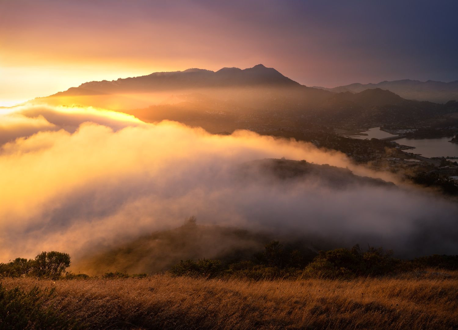 Fog rolls over golden hills at sunset with Mount Tamalpais silhouetted against an orange sky in Marin County, California.