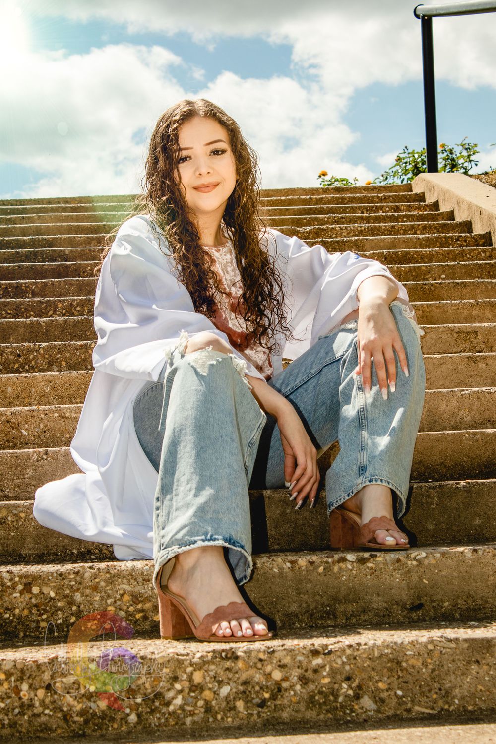 A person in a white blouse and jeans poses casually on concrete steps in warm sunlight.