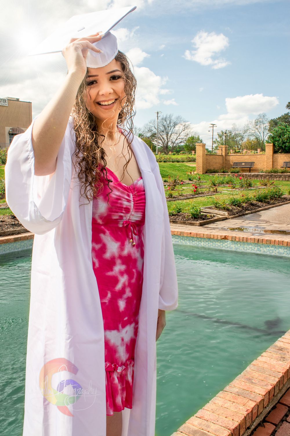 Graduate in white cap and gown with pink tie-dye dress poses by fountain during outdoor graduation photoshoot.