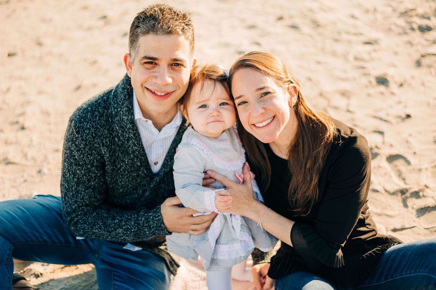 mother, father, and baby girl sitting in the sand at a beach
