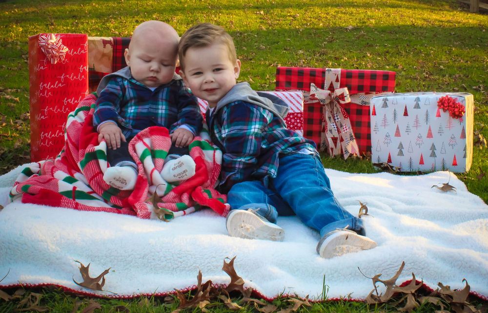 Two young children in plaid shirts sit on a blanket with Christmas presents outdoors.