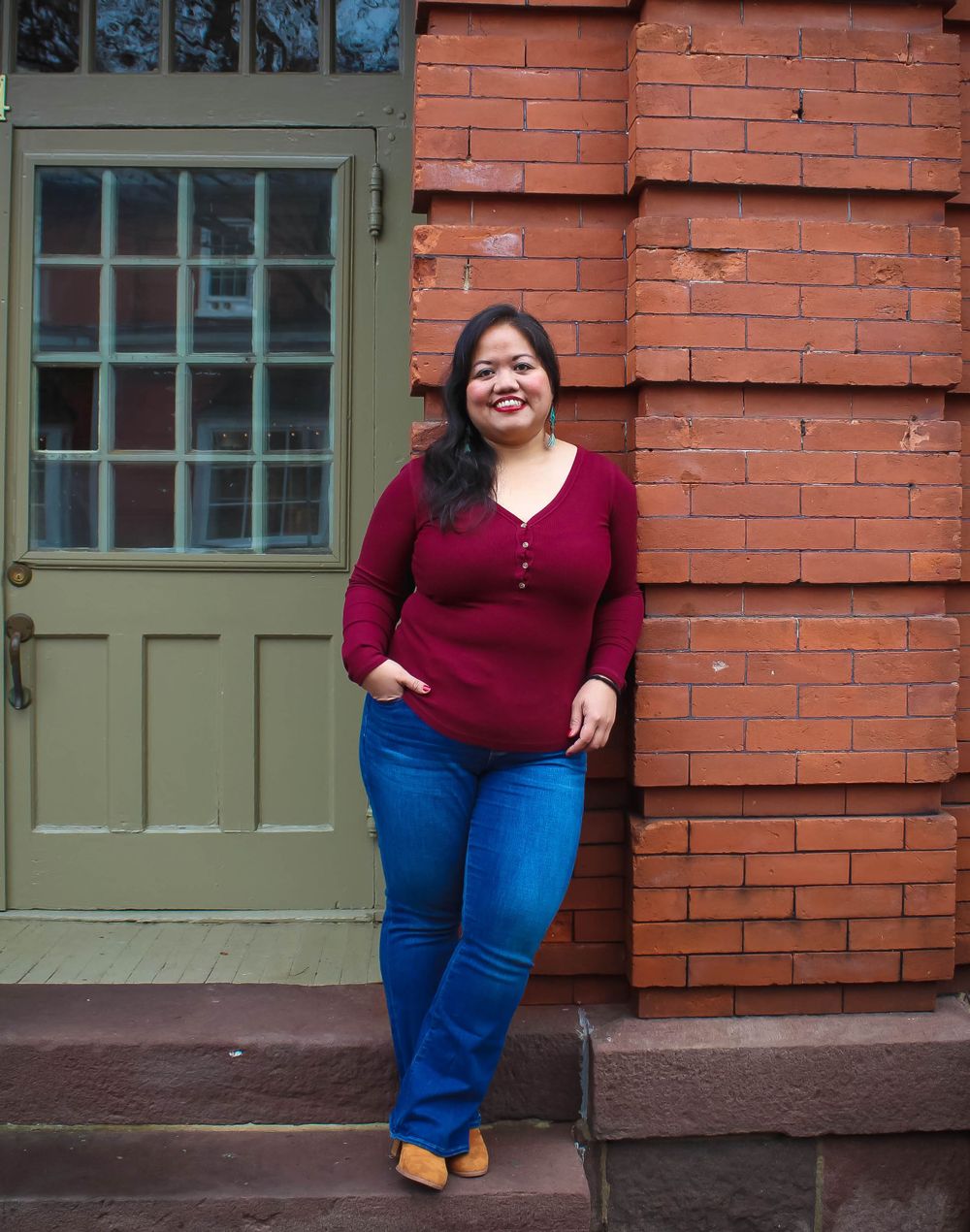 Woman in burgundy top and jeans poses casually against brick wall.