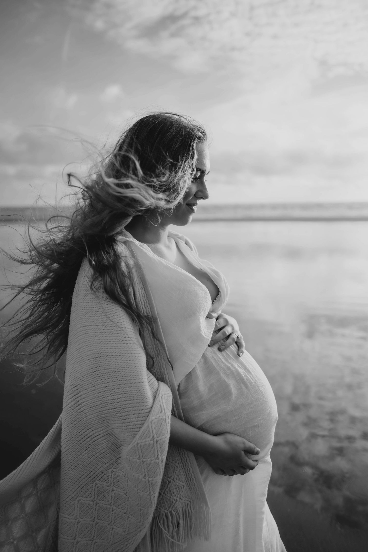 Artistic black and white photo of pregnant silhouette in flowing dress by water with wind-swept hair at sunset.