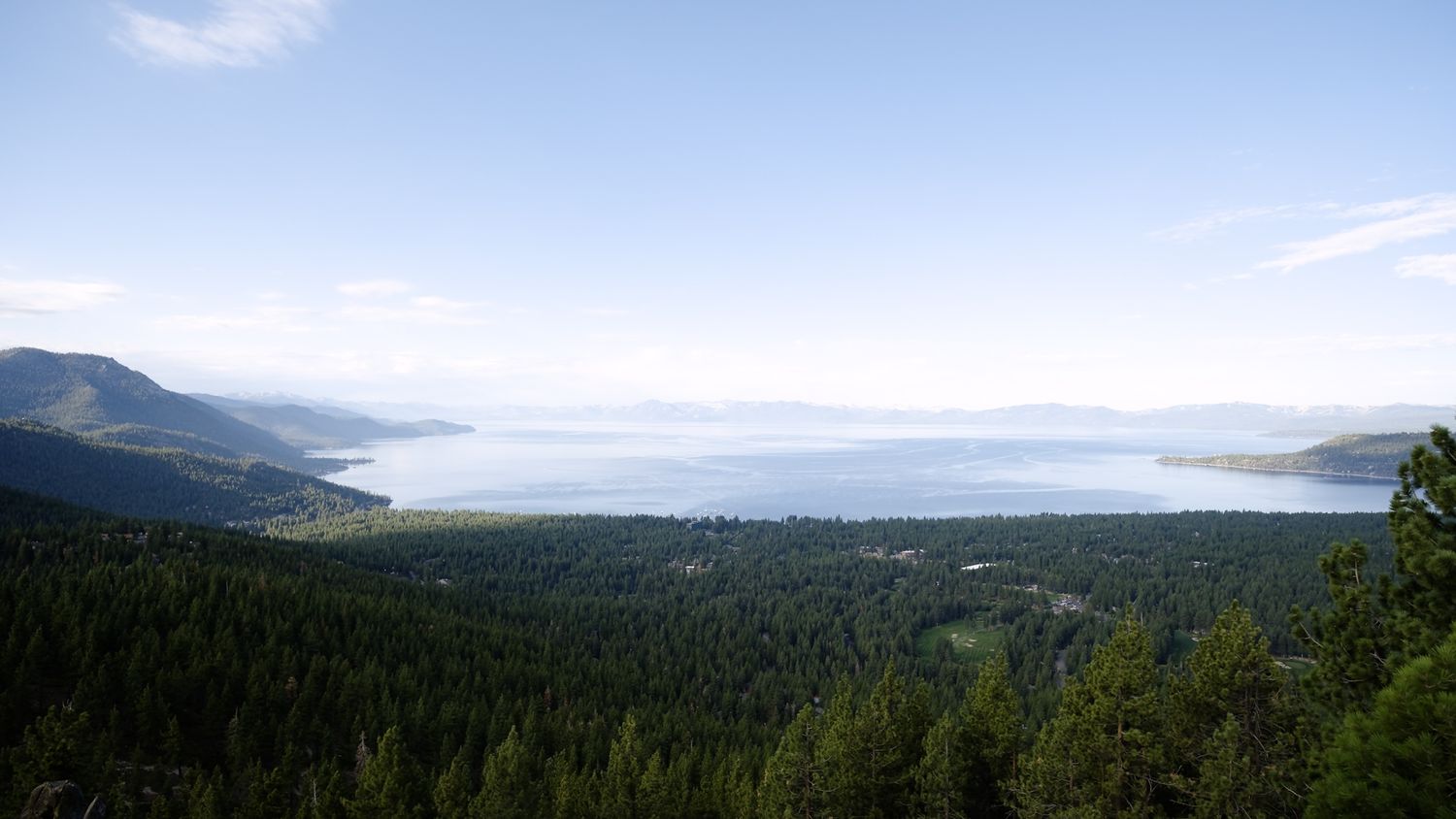 Aerial view of Lake Tahoe surrounded by dense forest and mountains under a blue sky and light cloud cover.