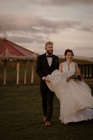 Wedding couple walking on grass with ornate red gazebo and moody sky in background.
