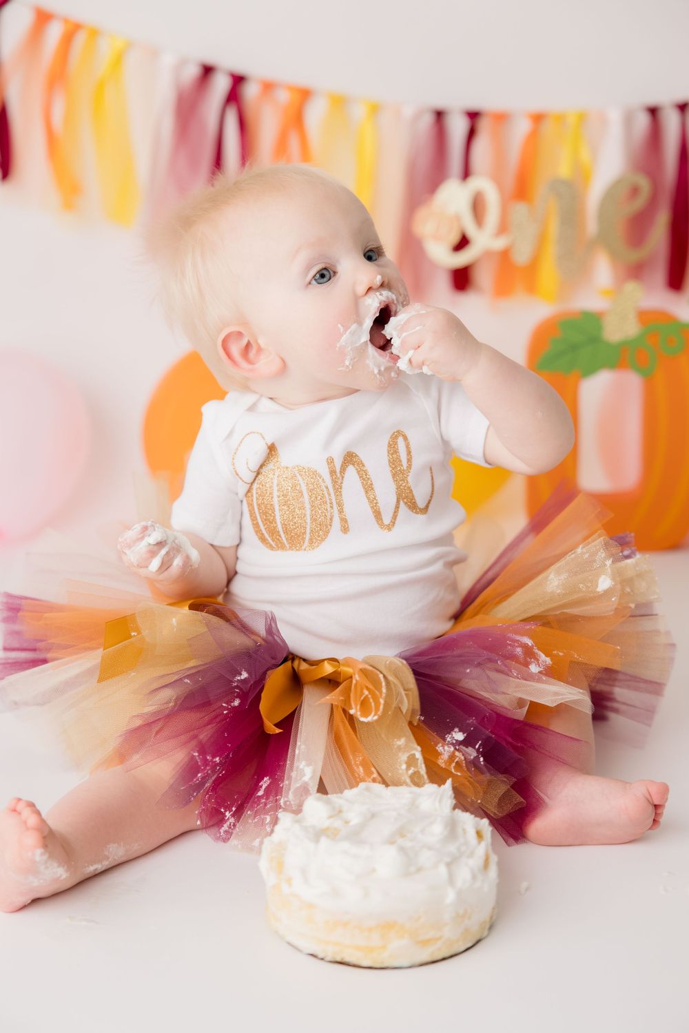 Baby wearing tutu and 'one' outfit sits with birthday cake against colorful party banner backdrop.