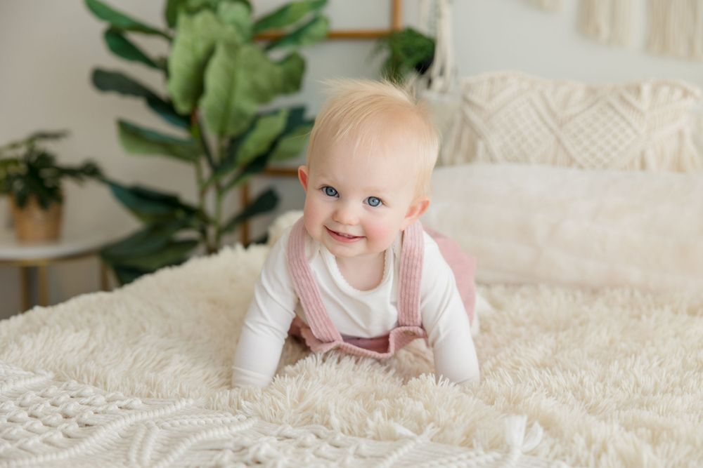 Baby wearing pink overalls crawls on white textured bedding with houseplants in background.