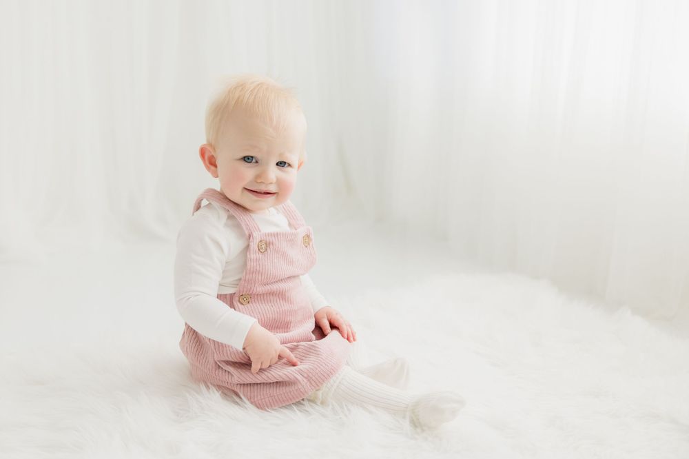 Baby in pink overalls and white shirt sitting on white blanket during portrait session.