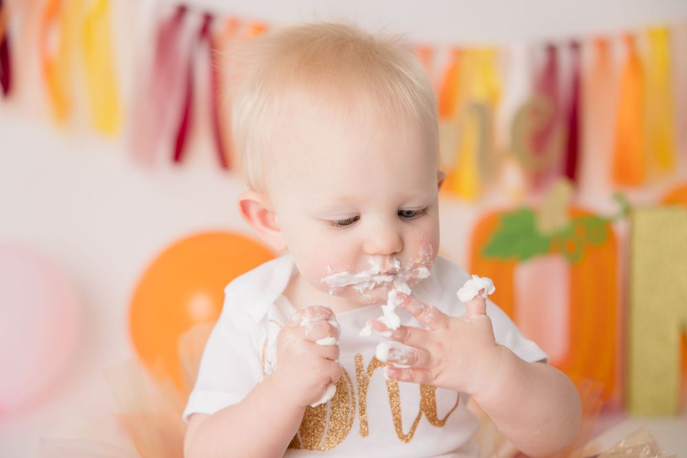 Baby in white and gold outfit enjoying cake during first birthday photoshoot with colorful backdrop.