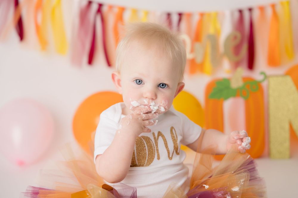 Baby wearing gold 'one' outfit sits among fall-colored decorations and bunting.