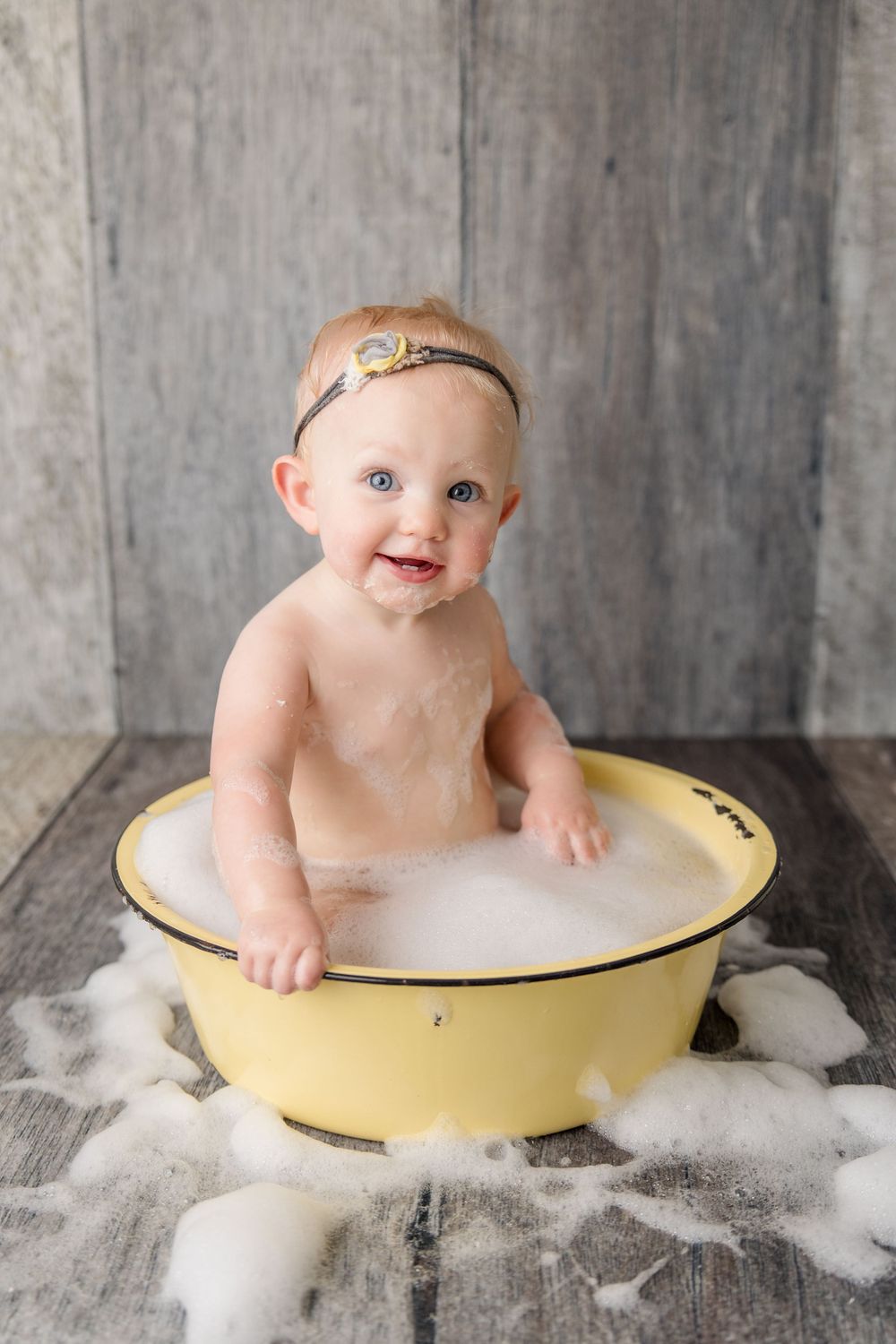 Baby sitting in yellow vintage washbasin surrounded by cotton-like bubbles on wood floor.