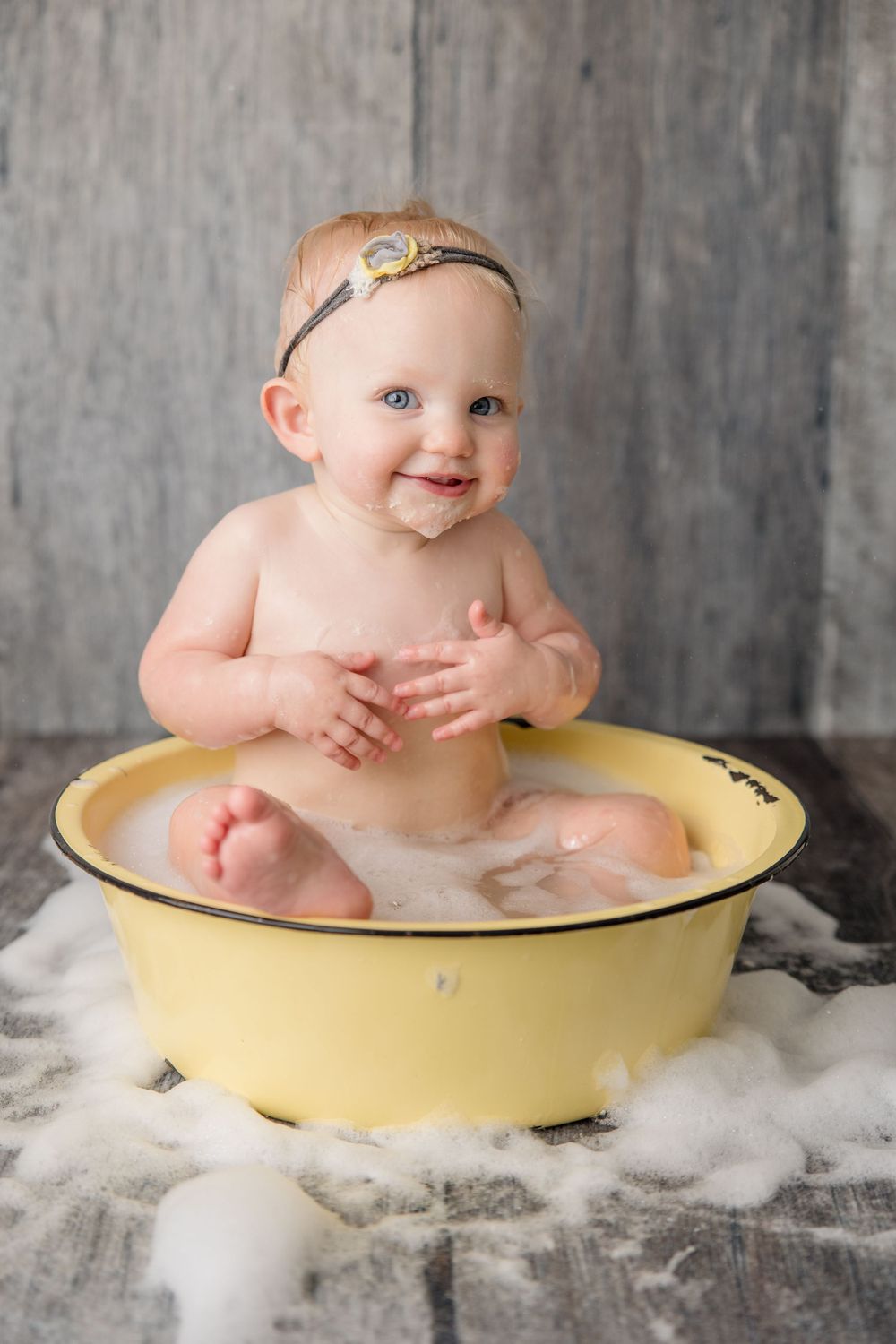 Baby sitting in yellow enamel basin during milk bath photoshoot on rustic wood floor.