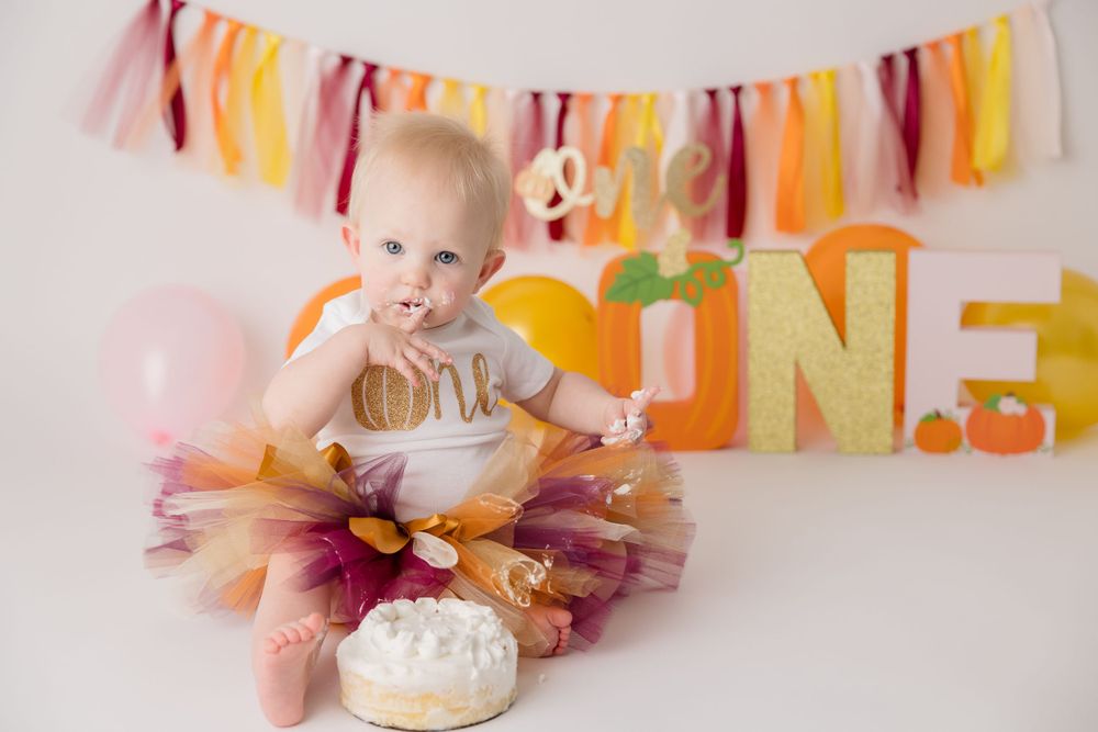 Baby in tulle dress reaches for birthday cake with festive banner and decorations behind.