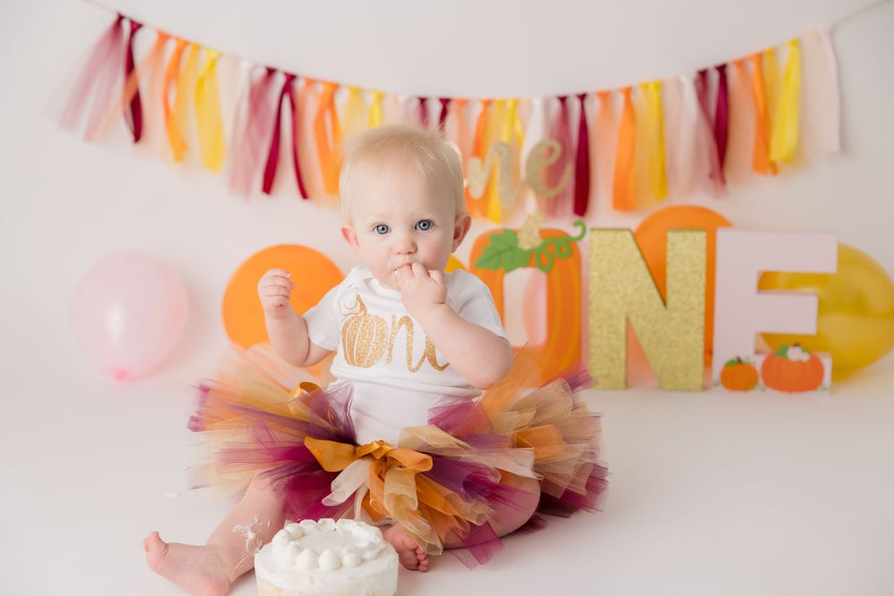 Baby in tutu dress celebrates first birthday with cake and colorful banner backdrop.