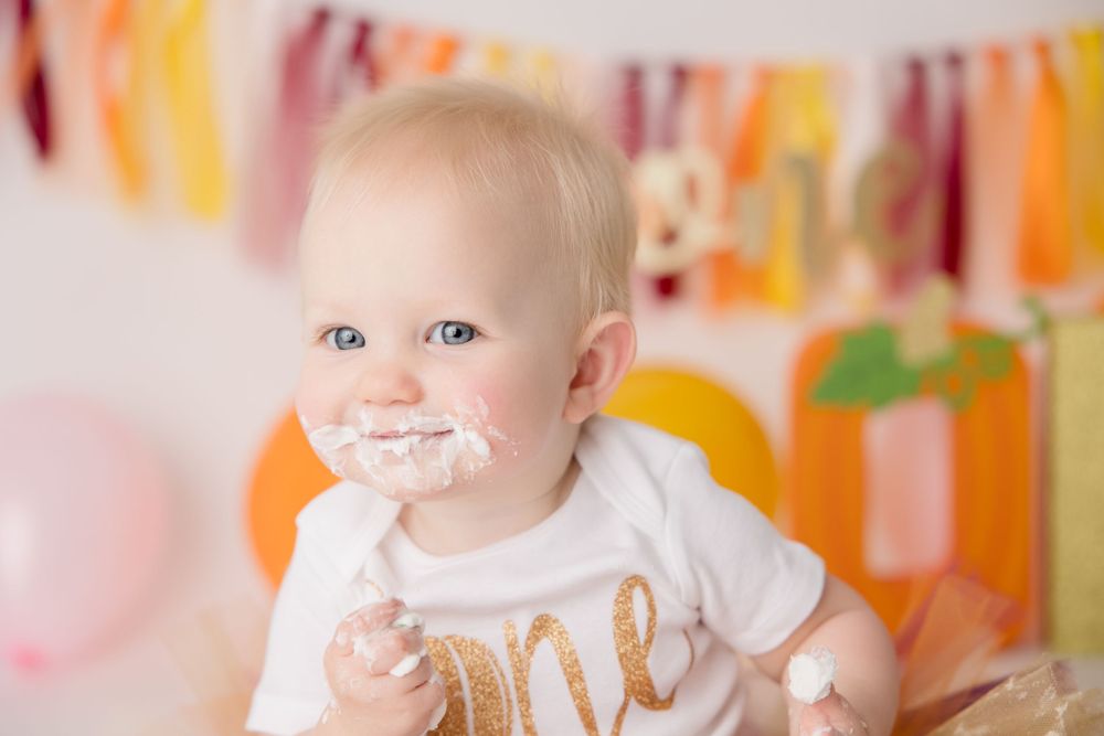 Baby with cake frosting on face sits in front of festive orange and pink bunting decorations.