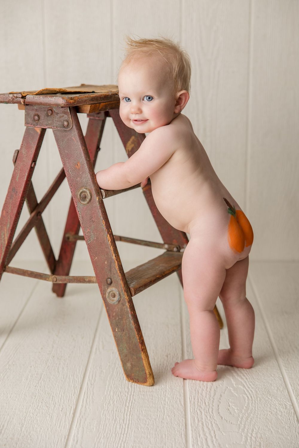 Baby in diaper stands holding rustic wooden ladder during photo shoot.