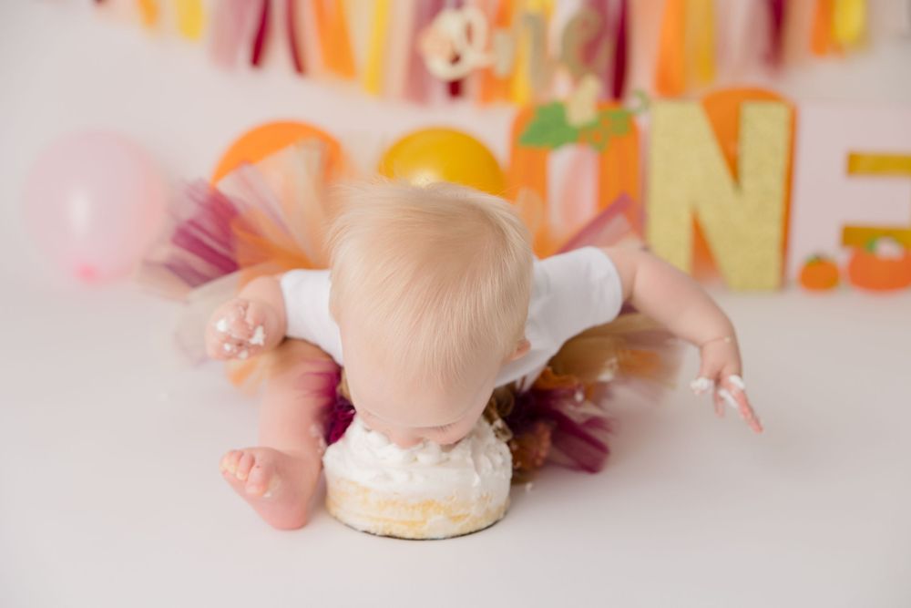 Baby in white outfit face down on floor with colorful bunting decorations overhead and small cake smash setup.