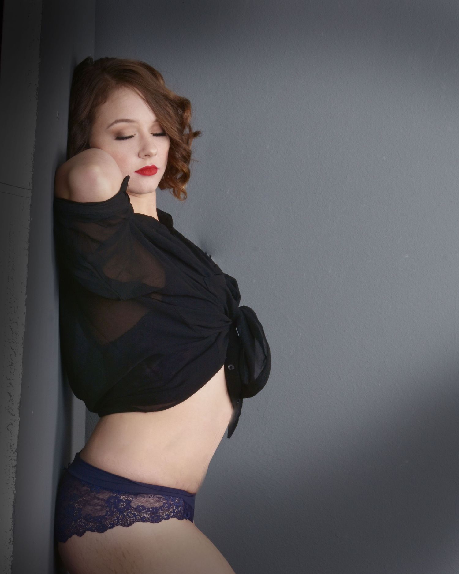 A boudoir portrait in monochrome lighting shows a figure in a black sheer top and navy lace underwear against a gray wall.