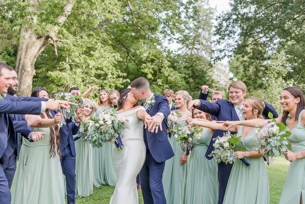 Wedding party laughing together outdoors with bridesmaids in sage green dresses holding white and purple bouquets.