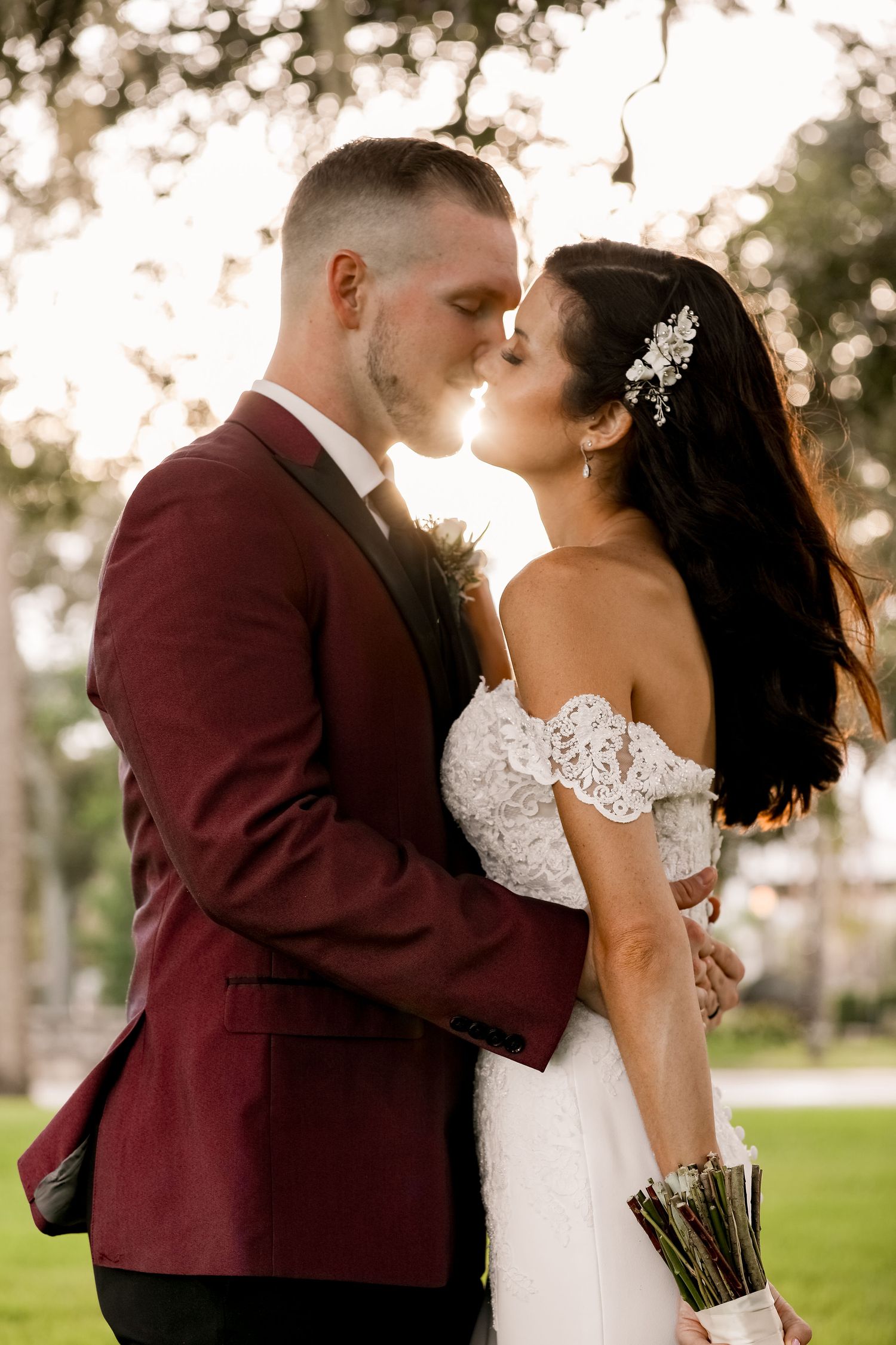 Romantic wedding couple shares a kiss outdoors in a garden setting at sunset, with burgundy suit and lace off-shoulder dress.