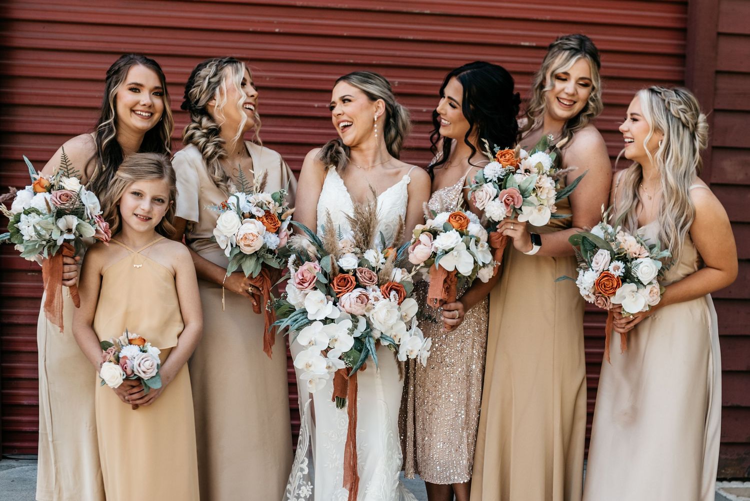 Wedding party in neutral dresses holding elegant white and pink floral bouquets against red barn wall backdrop.