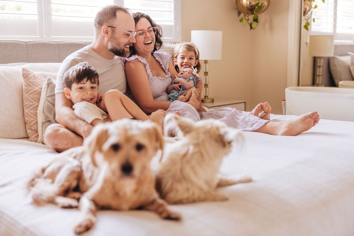 Family and dog relaxing together on white bedding in sunlit bedroom.