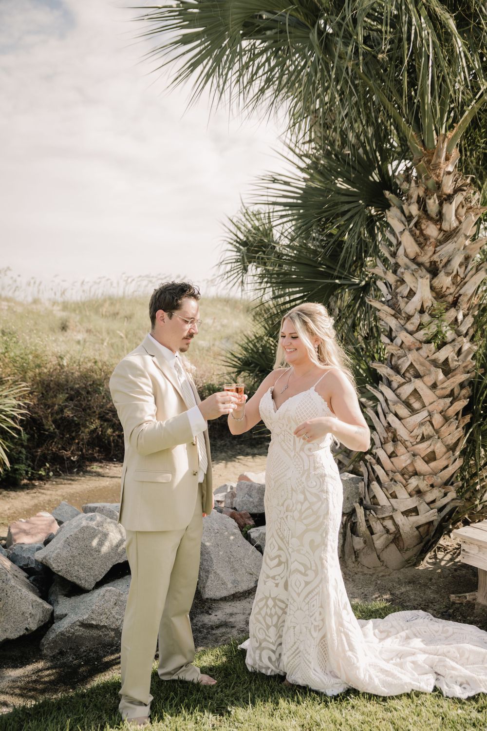 A wedding couple toasts and laughs together under a palm tree in an outdoor setting.