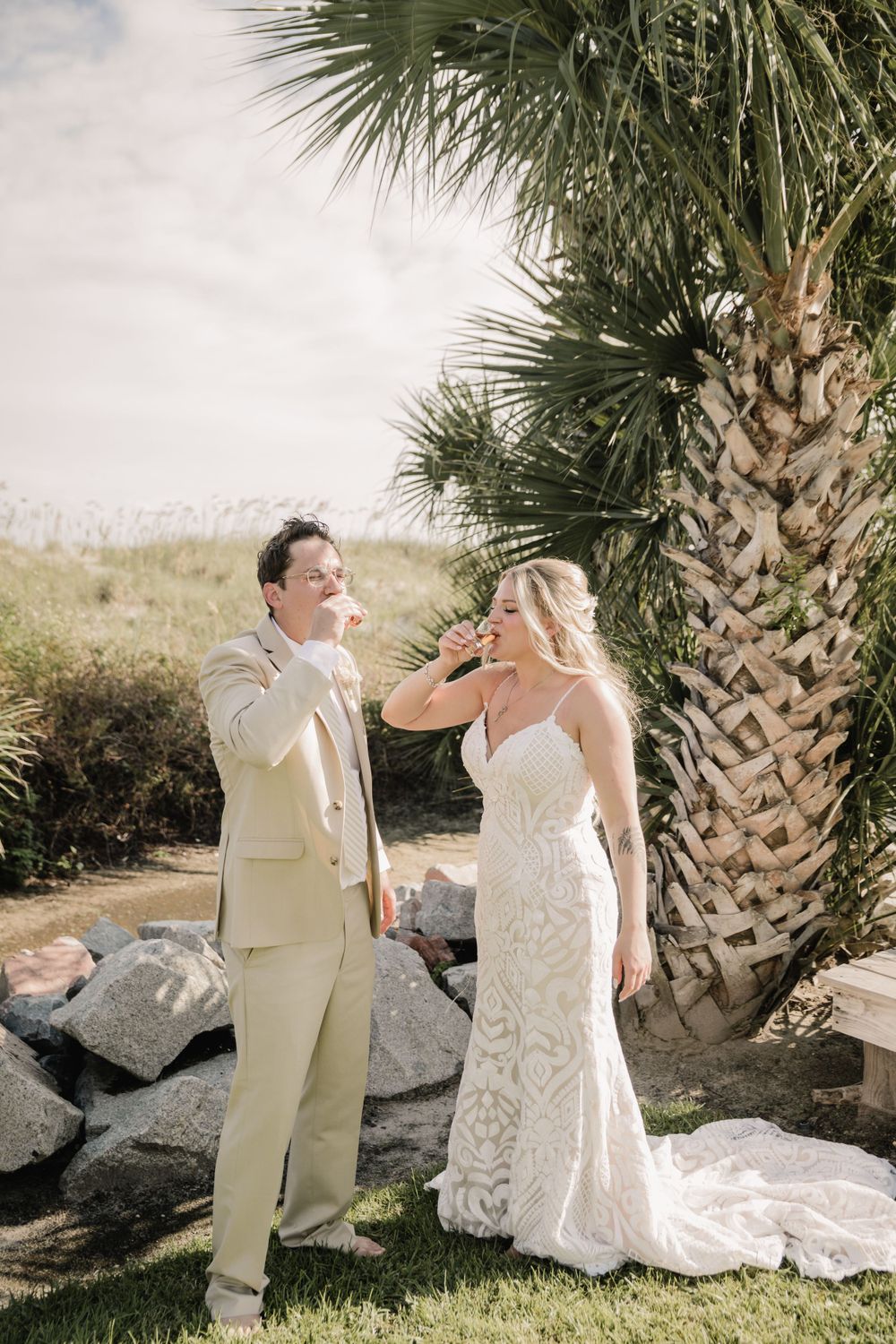 Newlyweds in light-colored attire share drinks during their tropical outdoor wedding celebration.
