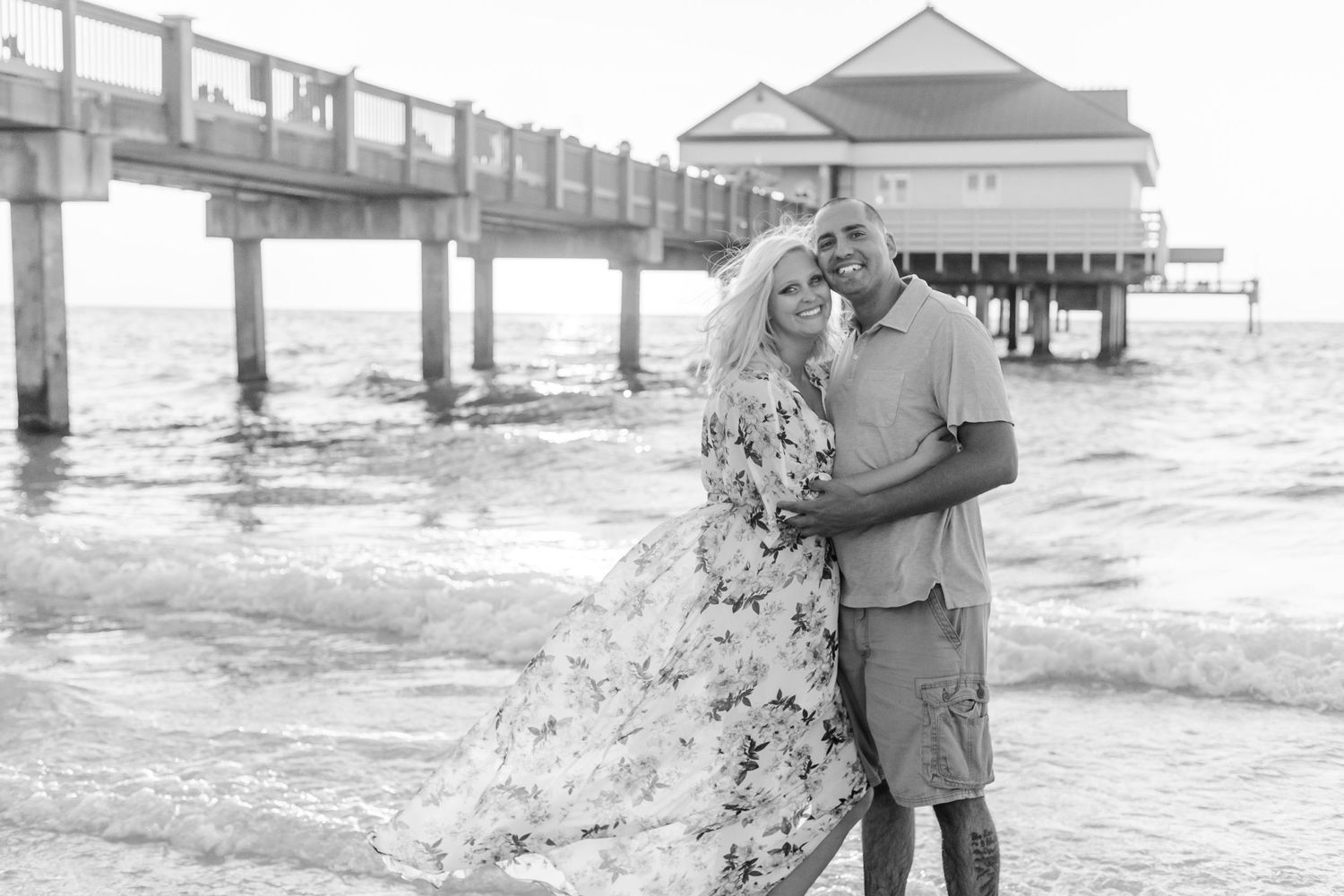 Black and white photo of couple embracing near beach pier at sunset.
