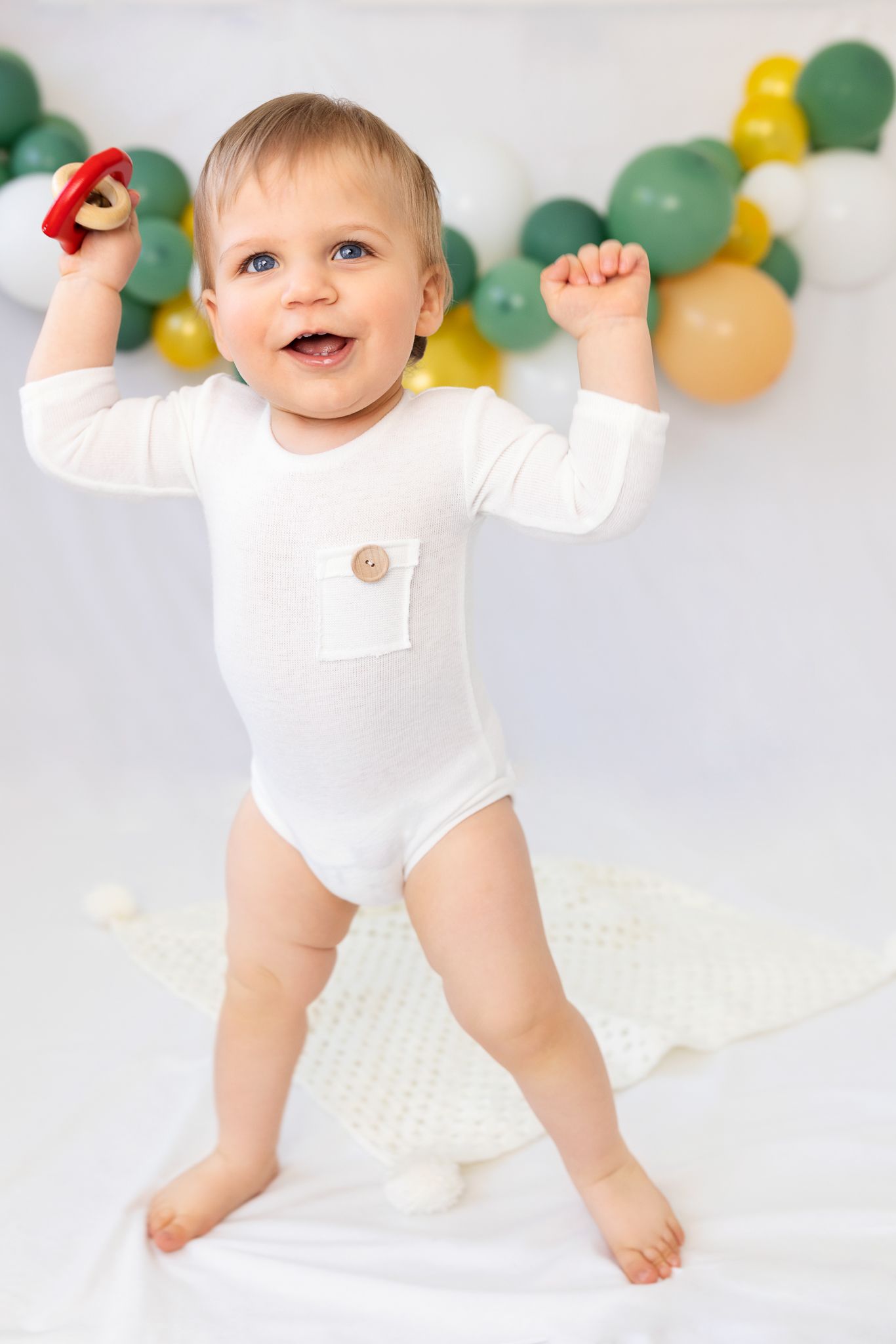 Happy baby in white onesie crawling on floor with green and yellow balloon decorations.