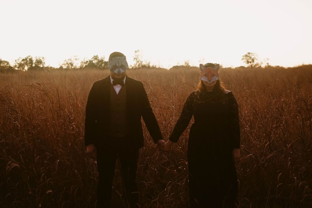 Two masked figures hold hands while walking through a field at dusk.