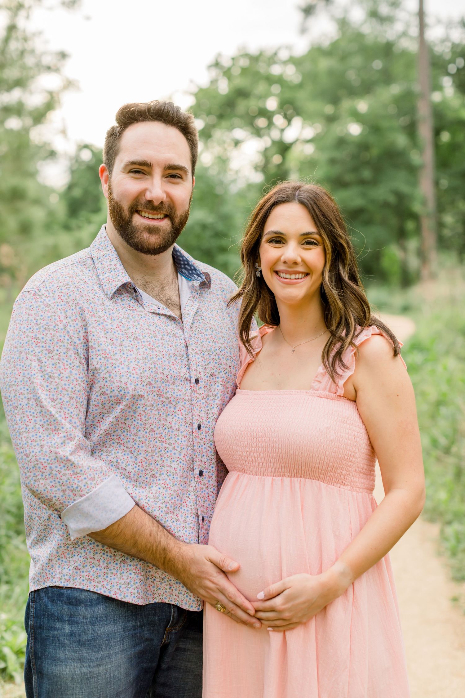 An expectant couple poses together on a wooded path wearing coordinating casual attire.