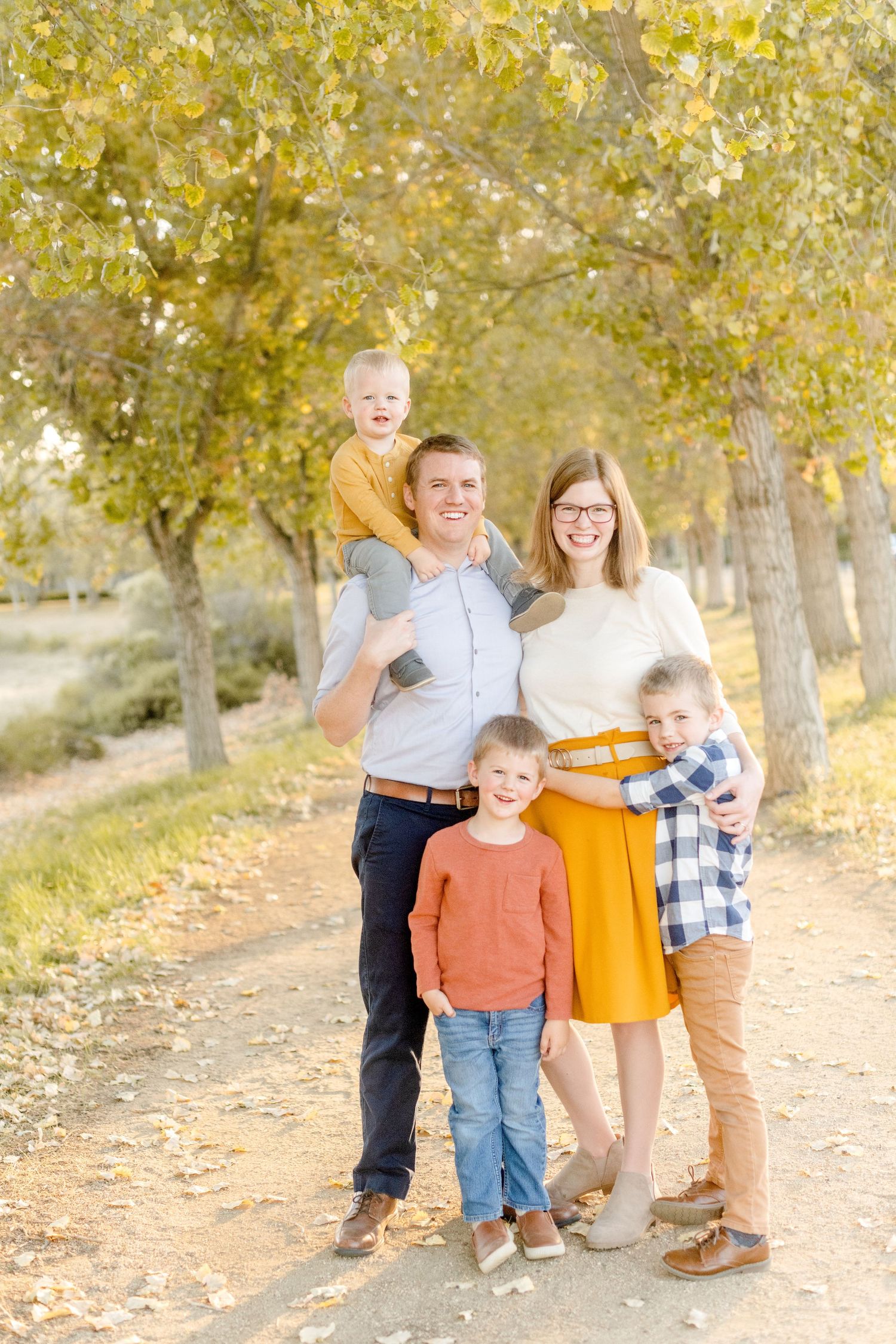 Family poses together on dirt path with golden autumn trees in background.