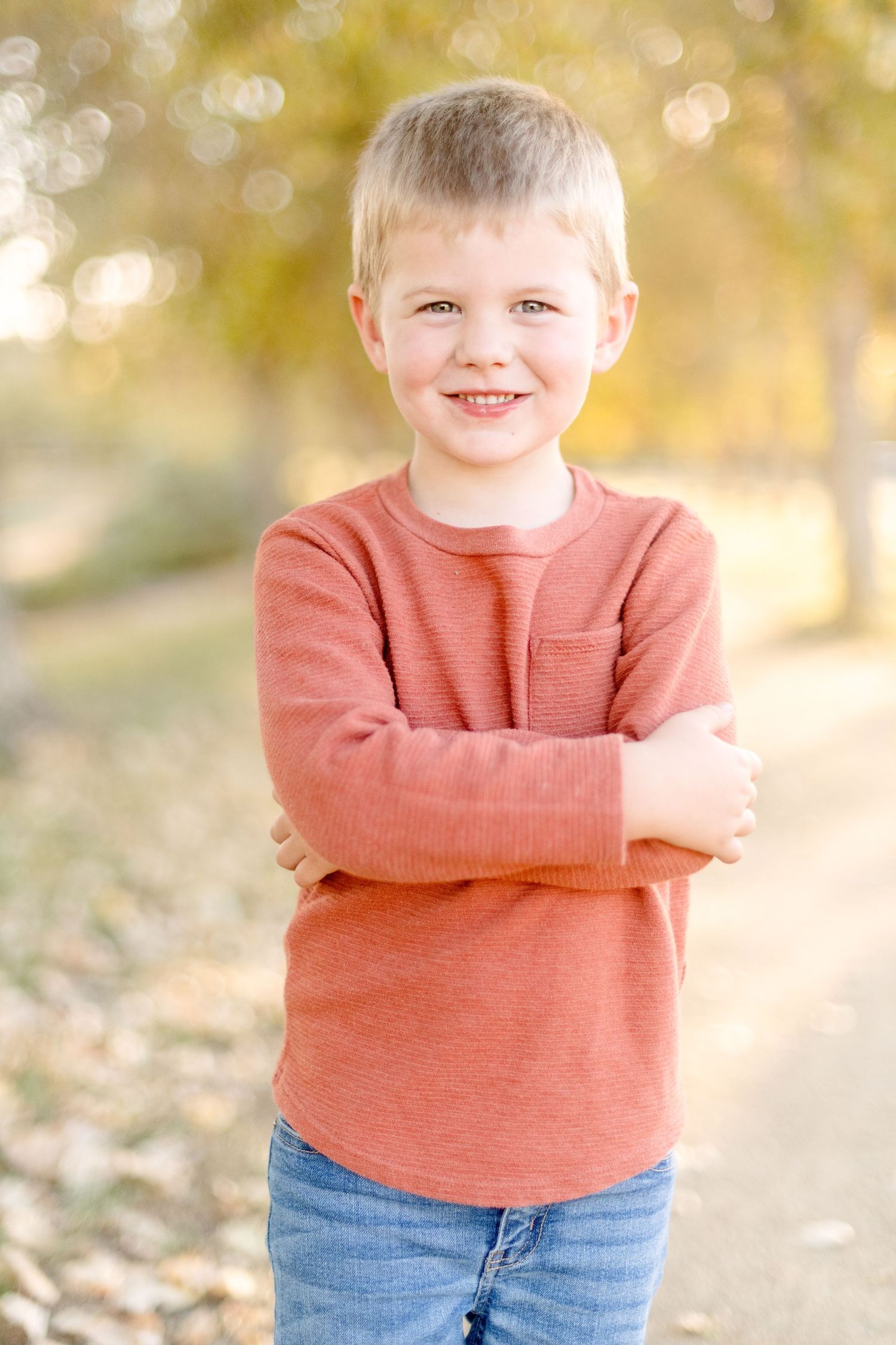 Child in coral colored sweater crosses arms and smiles outdoors with fall foliage in background.