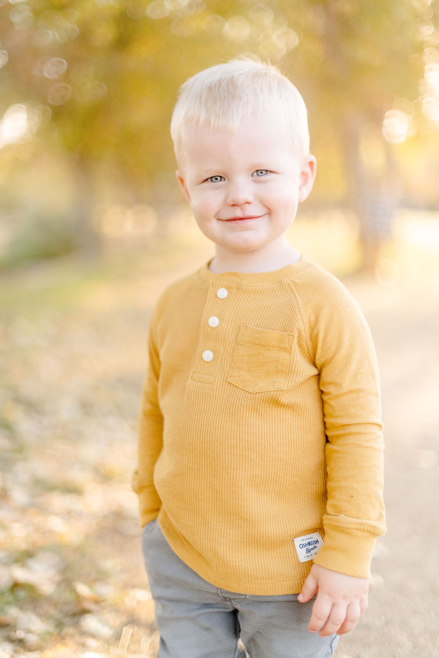 Small child in a yellow henley shirt smiles while standing outside on an autumn day.