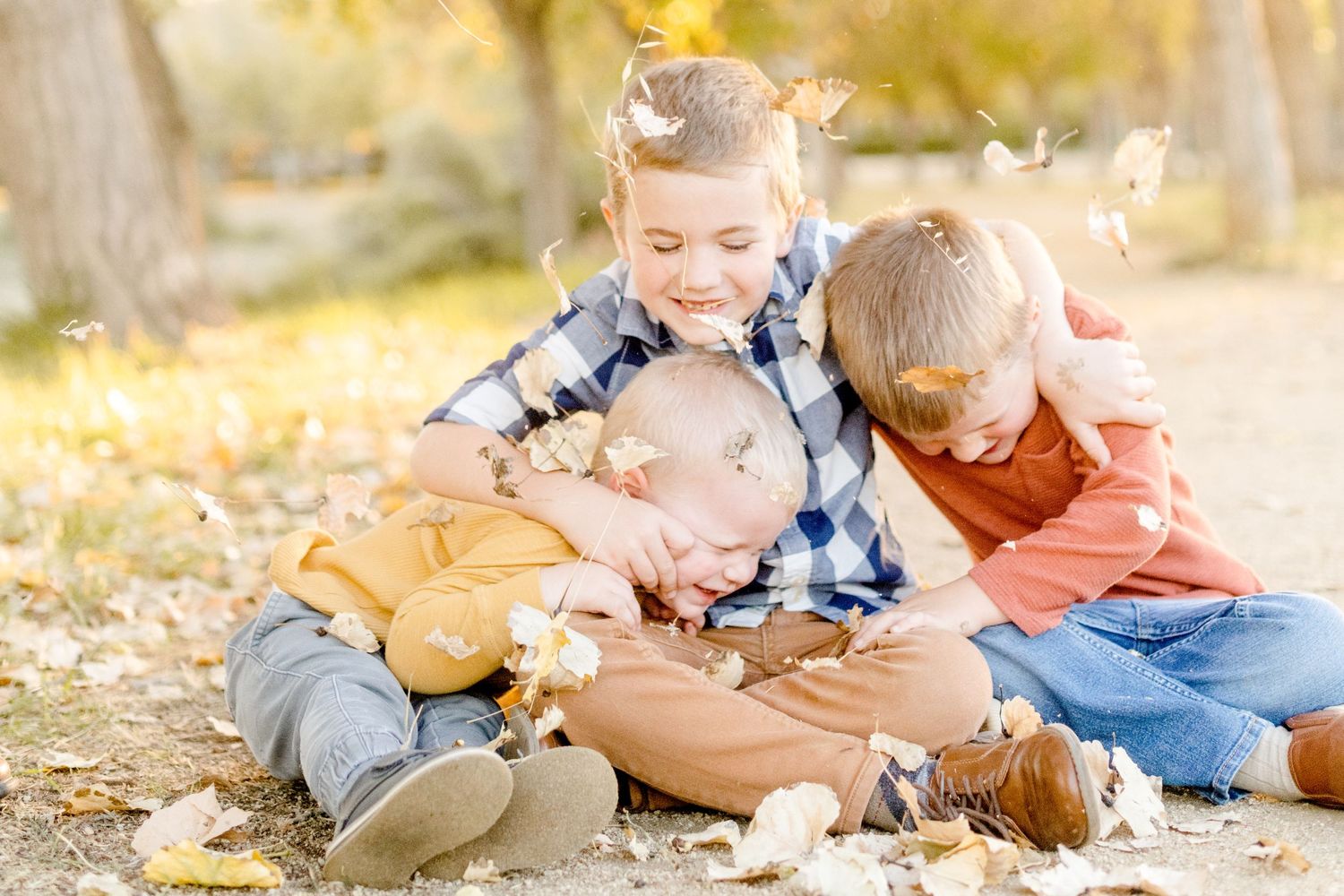 Three children lay together laughing on ground covered with fallen leaves.